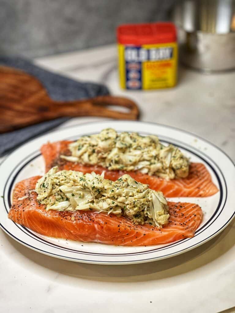 Two raw salmon filets that have been stuffed in the middle with a crabmeat mixture sitting on a white plate trimmed in blue.