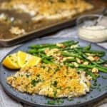 A piece of panko crusted baked haddock garnished with fresh parsley sitting on a blue plate with lemon wedges and green beans with sliced almonds.