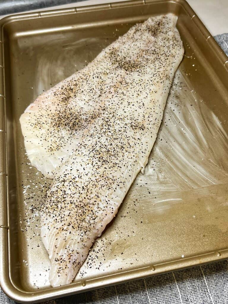 A large piece of haddock seasoned with salt and pepper sitting on a butter-lined baking sheet.