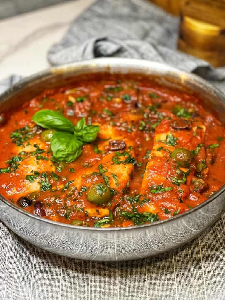 A deep sauté pan of three salmon filets in a red, puttanesca sauce.