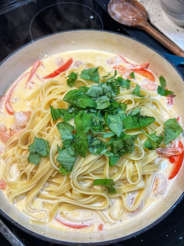 Al dente fettuccine pasta and fresh torn basil added to a pan of a creamy cayenne white sauce with red bell peppers.