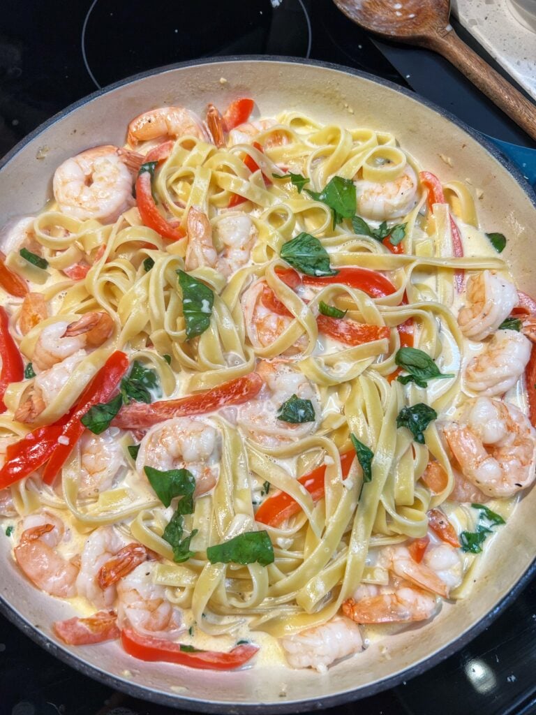 Pan of Creamy Cayenne Shrimp Pasta. Fettuccine is mixed in a creamy white sauce with cooked shrimp and red bell peppers. The dish is garnished with fresh torn basil.