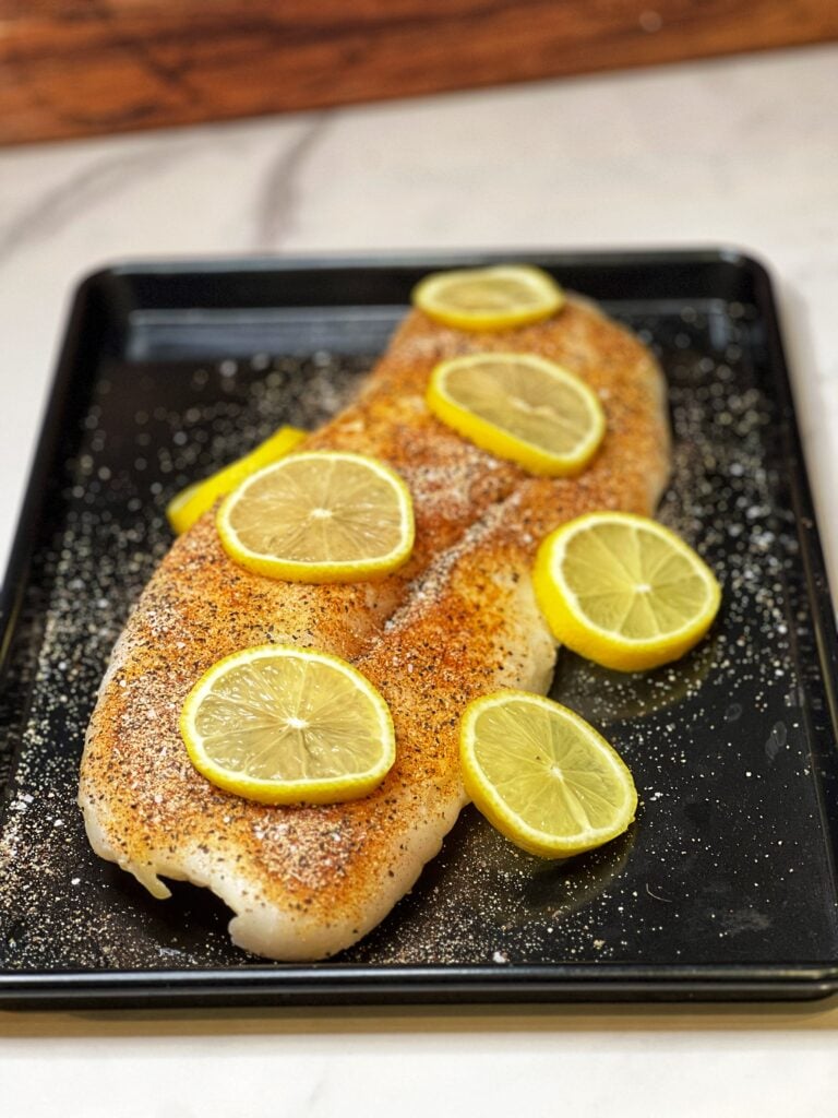 A large piece of uncooked, seasoned cod topped with a few lemon slices laying on a black sheet pan.