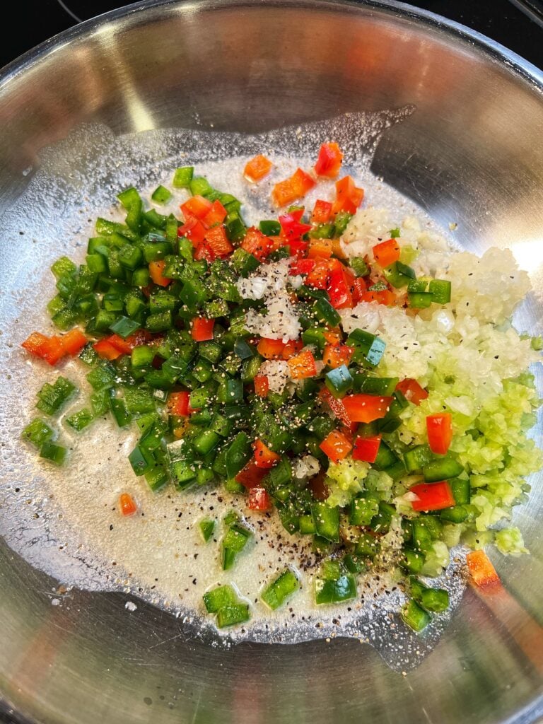 Diced red and green bell peppers, minced garlic, onions and celery sautéing in a stainless steel pan.