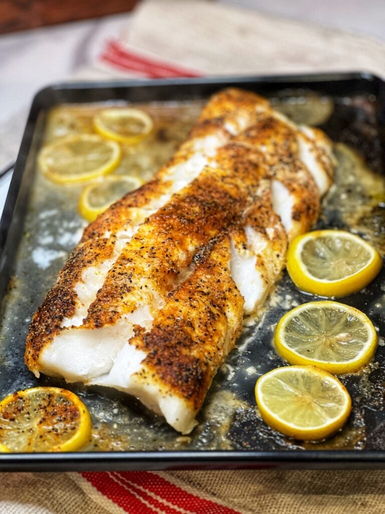 A large piece of seasoned baked cod with lemon slices sitting in a black sheet pan.