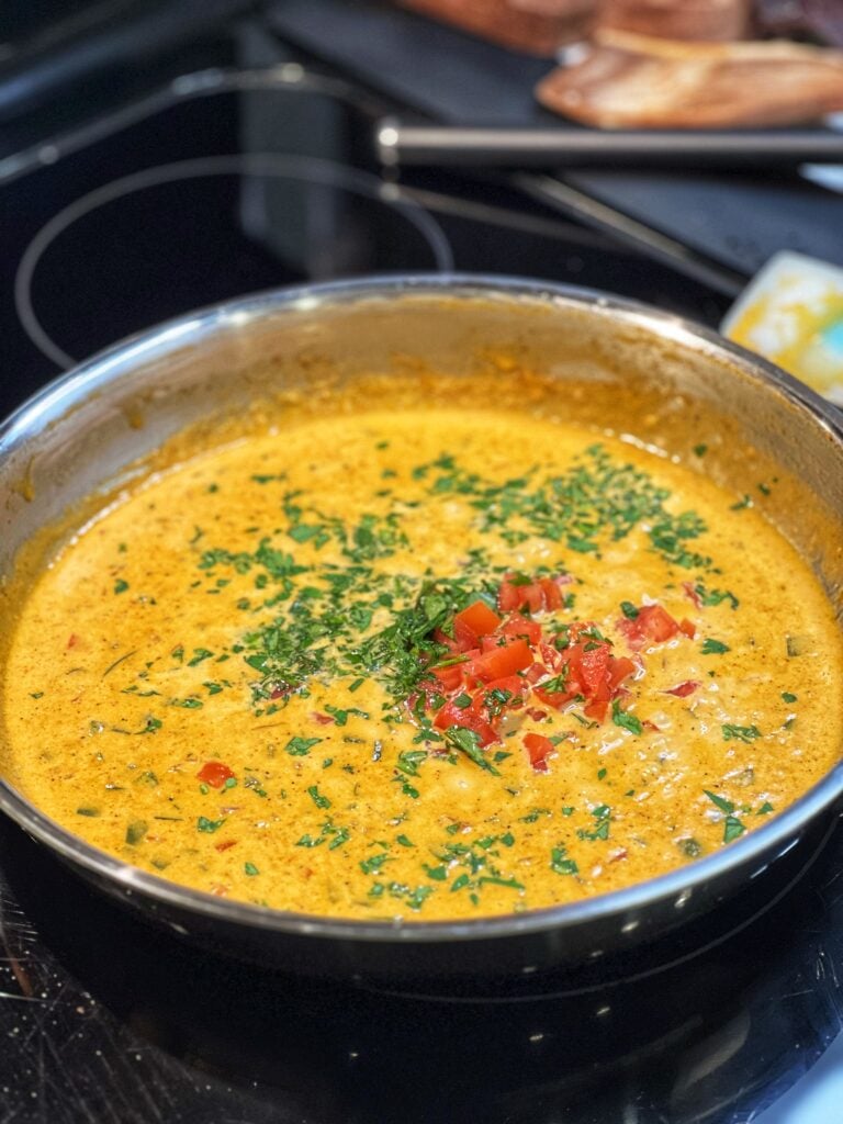 Creamy Creole sauce in a stainless steel pan with diced tomatoes and chopped parsley. The color of the sauce has a yellowish/orangish hue.