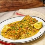 A large piece of baked cod on a white platter trimmed in black covered with a creamy Creole sauce. The sauce is a vibrant yellowish/orange and has diced tomatoes and green bell peppers.