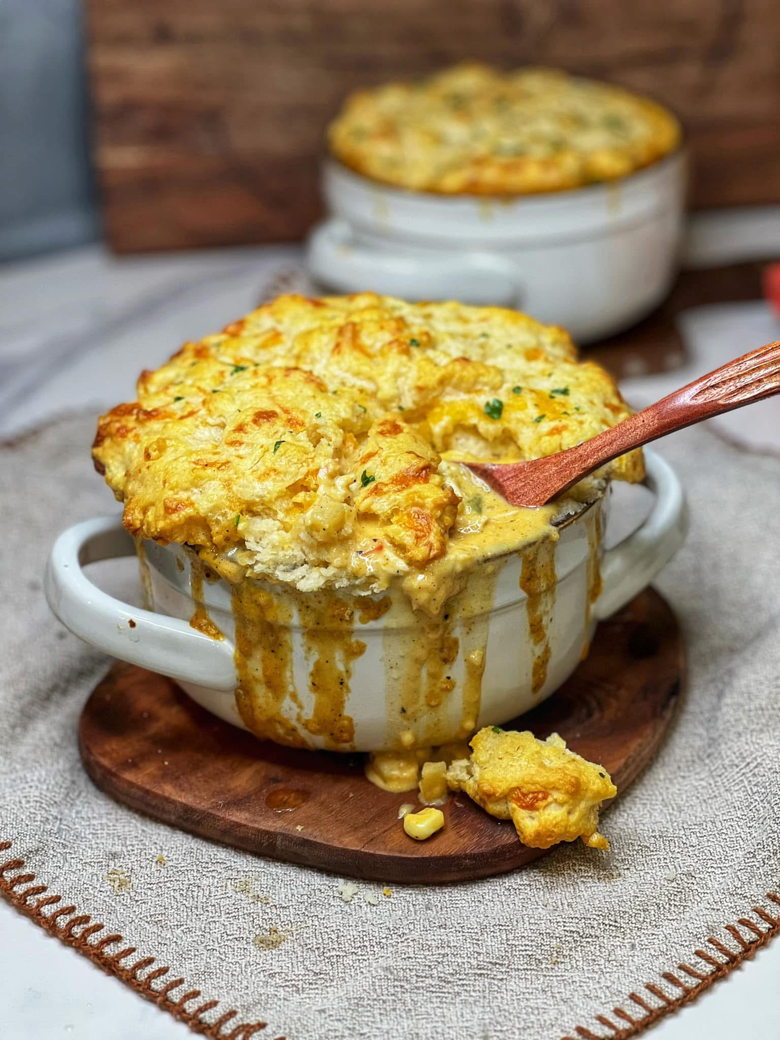 A white soup crock overflowing with Creole Chicken Pot Pie. The top crust of the pot pie is a baked, browned biscuit crust. There is a wooden spoon in the individual pie showing the inside contents. The bowl is sitting on a small wooden board.