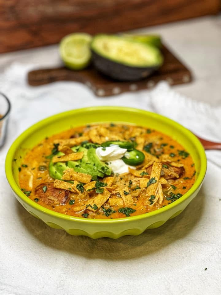 A green bowl filled with Creole chicken and rice soup. The soup has an orangish hue and is garnished with sliced avocado, a dollop of sour cream, crispy tortilla strips, sliced jalapeños and fresh chopped parsley.