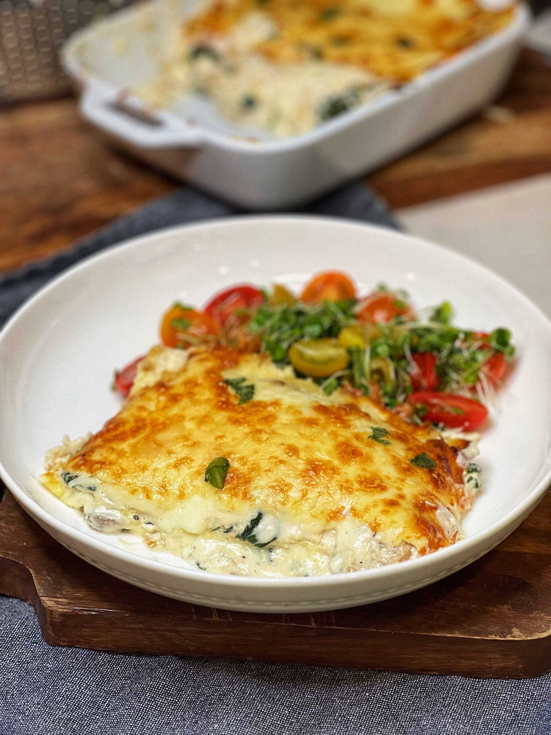 A piece of spinach and mushroom lasagna in a white bowl with a cherry tomato salad behind the lasagna.