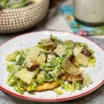 A white plate trimmed in orange holding a pounded down chicken breast topped with a Creamy Caesar Salad. The salad is made with chopped Romaine lettuce, homemade brown croutons and creamy dressing. The salad is garnished with shaved Parmesan and cracked black pepper.
