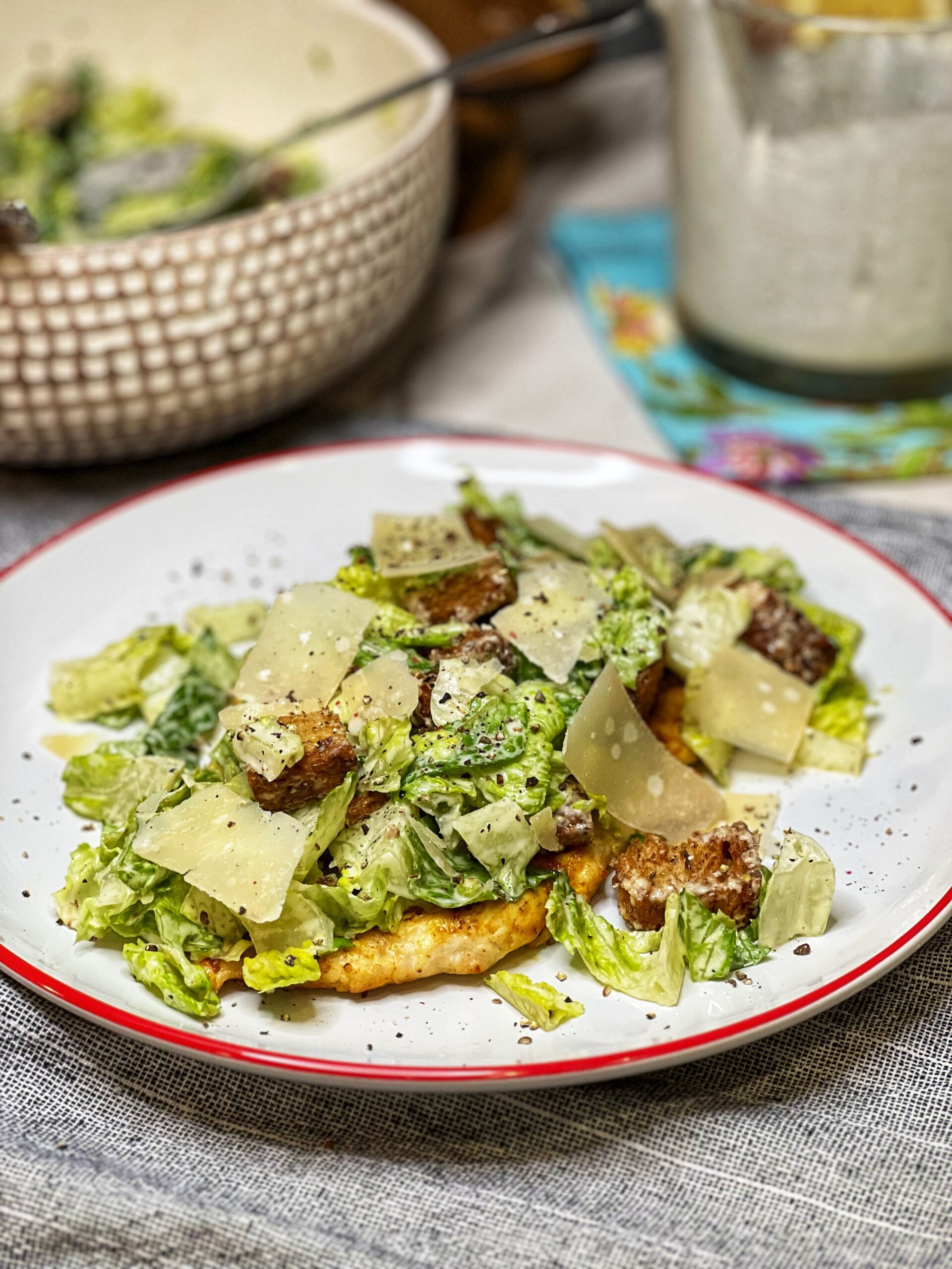A white plate trimmed in orange holding a pounded down chicken breast topped with a Creamy Caesar Salad. The salad is made with chopped Romaine lettuce, homemade brown croutons and creamy dressing. The salad is garnished with shaved Parmesan and cracked black pepper.
