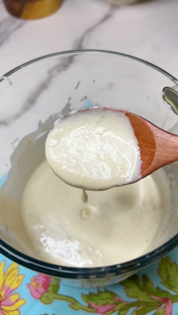 A wooden spoon holding creamy Caesar salad dressing over a jar of the dressing. The dressing is off-white in color.