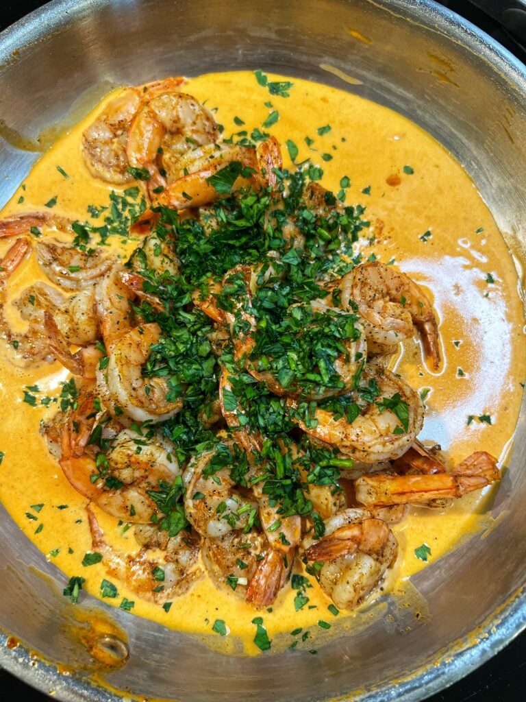 Seared shrimp and chopped parsley added to a pan of Cajun cream sauce. The sauce is orangish in color.