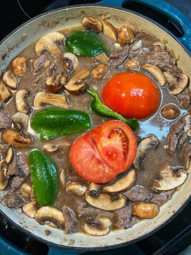 A pan of brown gravy with strips of steak, sliced mushrooms, green pepper strips and a halved tomato.