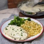 A round white plate with orange trim holding a deconstructed chicken cordon bleu covered in a mustard cream sauce with buttered noodles and steamed broccoli on the side.