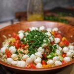 A large wooden bowl with unmixed ingredients for a chicken orzo pasta salad with cherry tomatoes, cucumbers, mozzarella and fresh herbs.