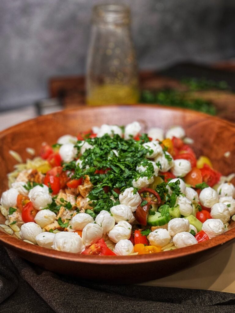 A large wooden bowl with unmixed ingredients for a chicken orzo pasta salad with cherry tomatoes, cucumbers, mozzarella and fresh herbs.