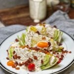 A classic iceberg wedge salad. Three wedges of iceberg lettuce on a white platter topped with halved cherry tomatoes, thinly sliced red onion, crisp bacon pieces and homemade blue cheese dressing.