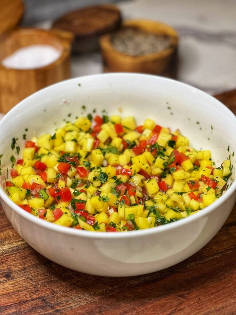 A white bowl of mango salsa. The salsa has diced mango, diced red bell pepper, diced tomato, and fresh cilantro.
