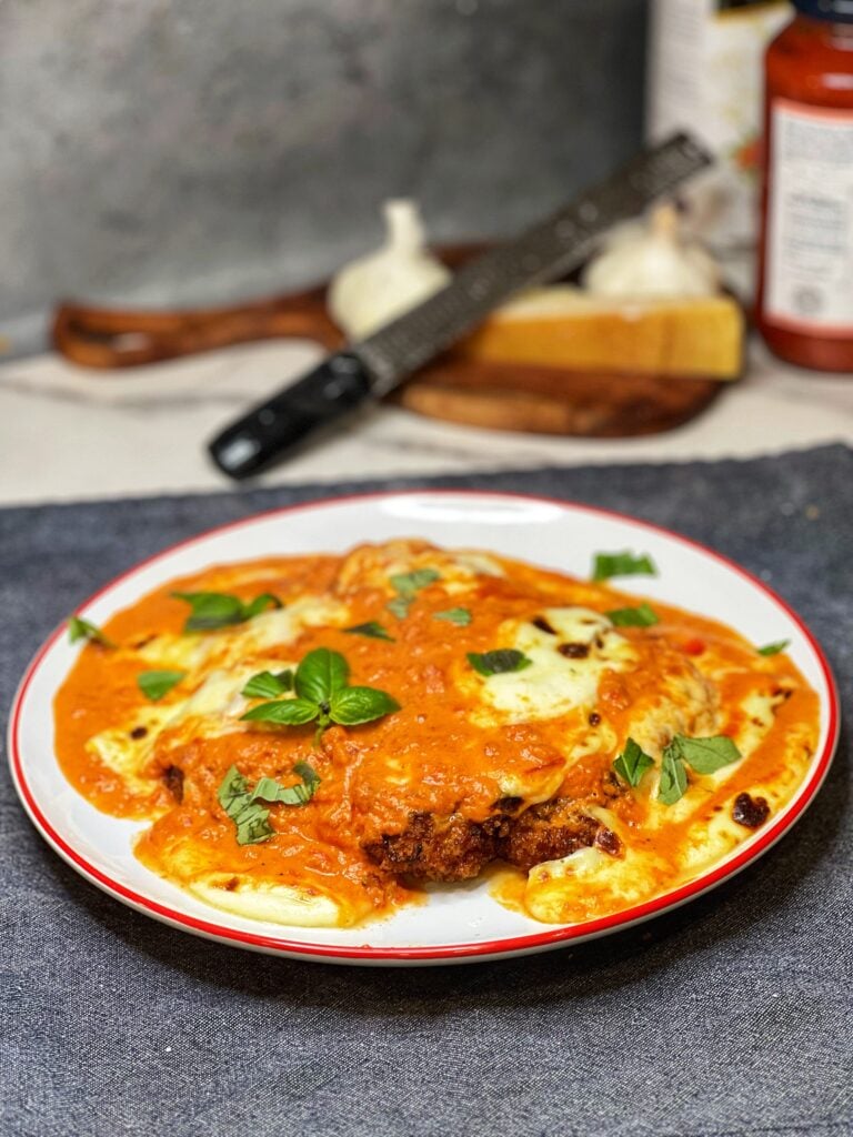 A piece of chicken Parmesan alla vodka on a white plate. A piece of crispy chicken is covered in a orangish sauce that had melted mozzarella on top. The dish is garnished with fresh basil.