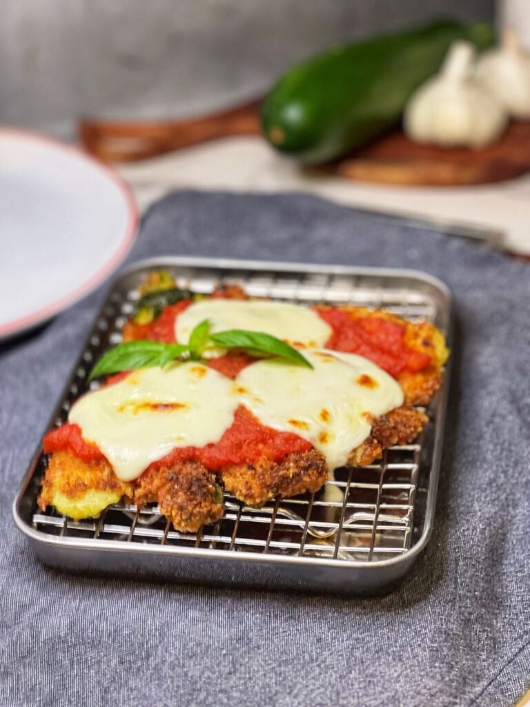 A piece of crispy fanned zucchini Parmesan sitting on a small pan with a wire rack. The zucchini has been breaded, fried then topped with marinara sauce and fresh mozzarella. It was then placed under the broiler to melt and slightly brown the cheese.