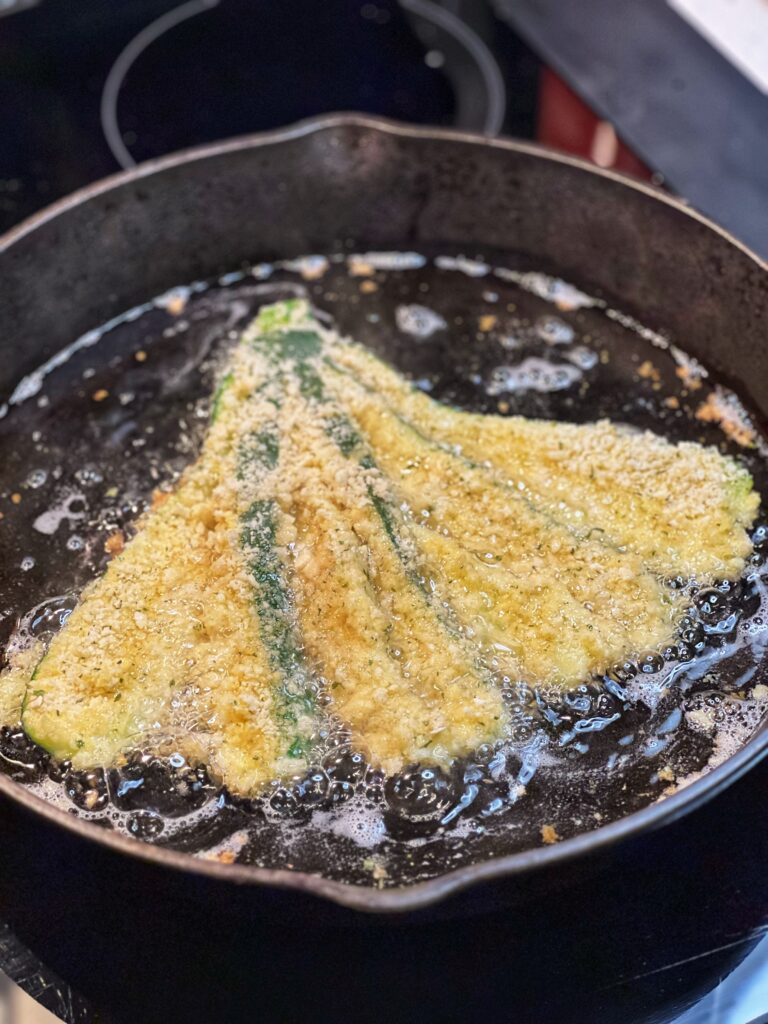 A fanned zucchini that has been coated in Panko breadcrumbs shallow frying in a cast iron pan.