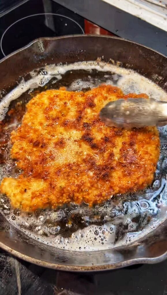A piece of breaded chicken frying in a cast iron pan for chicken Parmesan alla vodka.