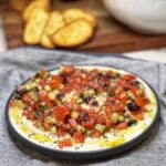A shallow blue plate filled with a whipped feta tomato salad. Whipped feta is the base for a tiny diced tomato salad with cucumber, red onion and Kalamata olives.