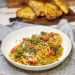 A white bowl filled with one pan pasta puttanesca. It's spaghetti covered in a sauce of cooked cherry tomatoes, olives, capers, garlic and fresh herbs. The dish is garnished with Parmesan cheese.