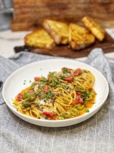 A white bowl filled with one pan pasta puttanesca. It's spaghetti covered in a sauce of cooked cherry tomatoes, olives, capers, garlic and fresh herbs. The dish is garnished with Parmesan cheese.