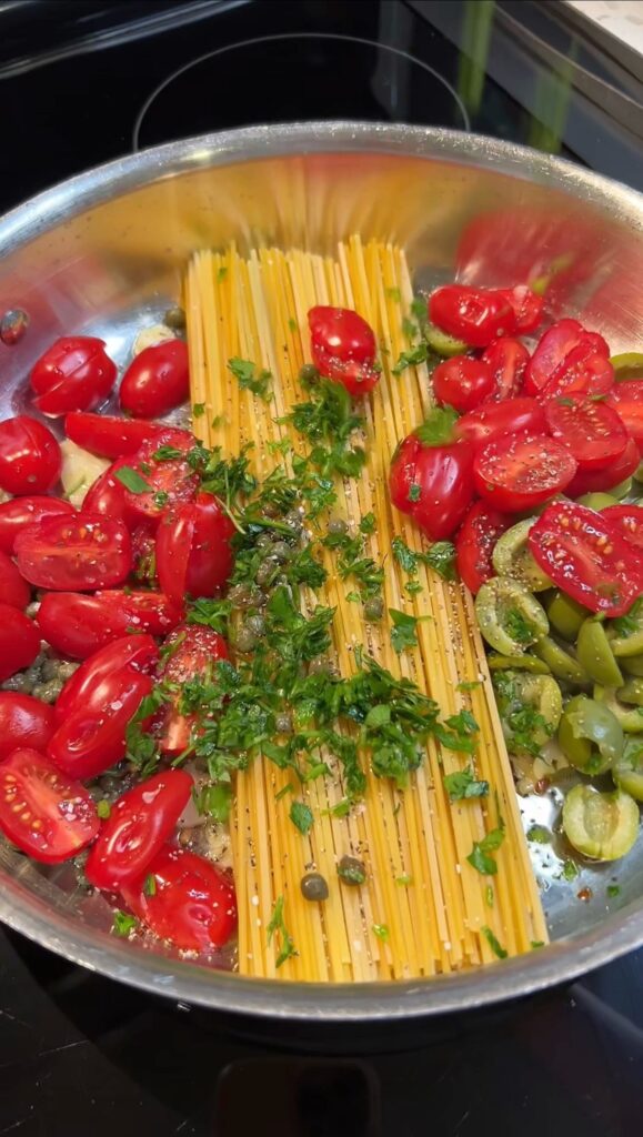 A deep sauté pan filled with dried spaghetti, halved cherry tomatoes, fresh parsley, olives, capers and thinly sliced garlic.
