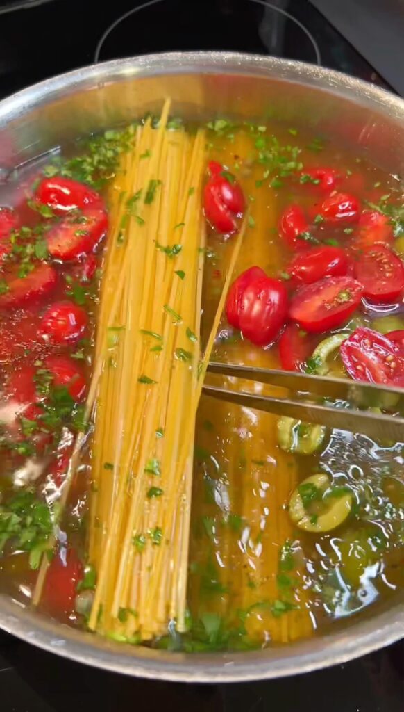 Chicken stock in a pan with dried pasta, cherry tomatoes, olives, capers, sliced garlic and parsley for a One Pan Pasta Puttanesca.
