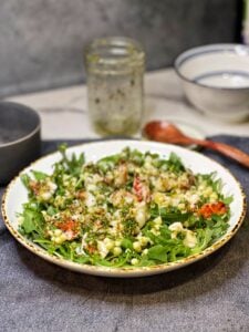 A white bowl trimmed in gold filled with a Lobster Corn Salad. The base of the salad is baby arugula topped with chunks of cooked lobster tail meat and fresh corn.