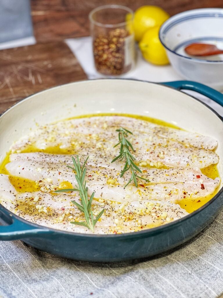 A blue enameled cast iron casserole holding three pieces of uncooked fish topped with a citrus sauce and two sprigs of rosemary.