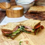 An argula BLT sandwich cut in half and sitting on a piece of parchment paper. You can see the tomato, arugula,bacon, sliced avocado and mayo from an inside view.