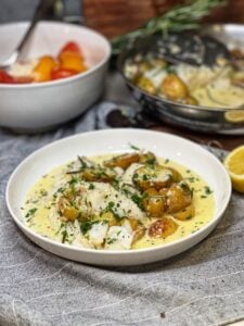 A white bowl filled with cod with a rosemary cream sauce and roasted potatoes. The dish is garnished with fresh chopped parsley.