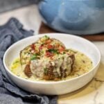 Creamy canned clam chowder poured on top of a baked potato that is sitting in a white bowl. The dish is garnished with crisp bacon and fresh parsley.