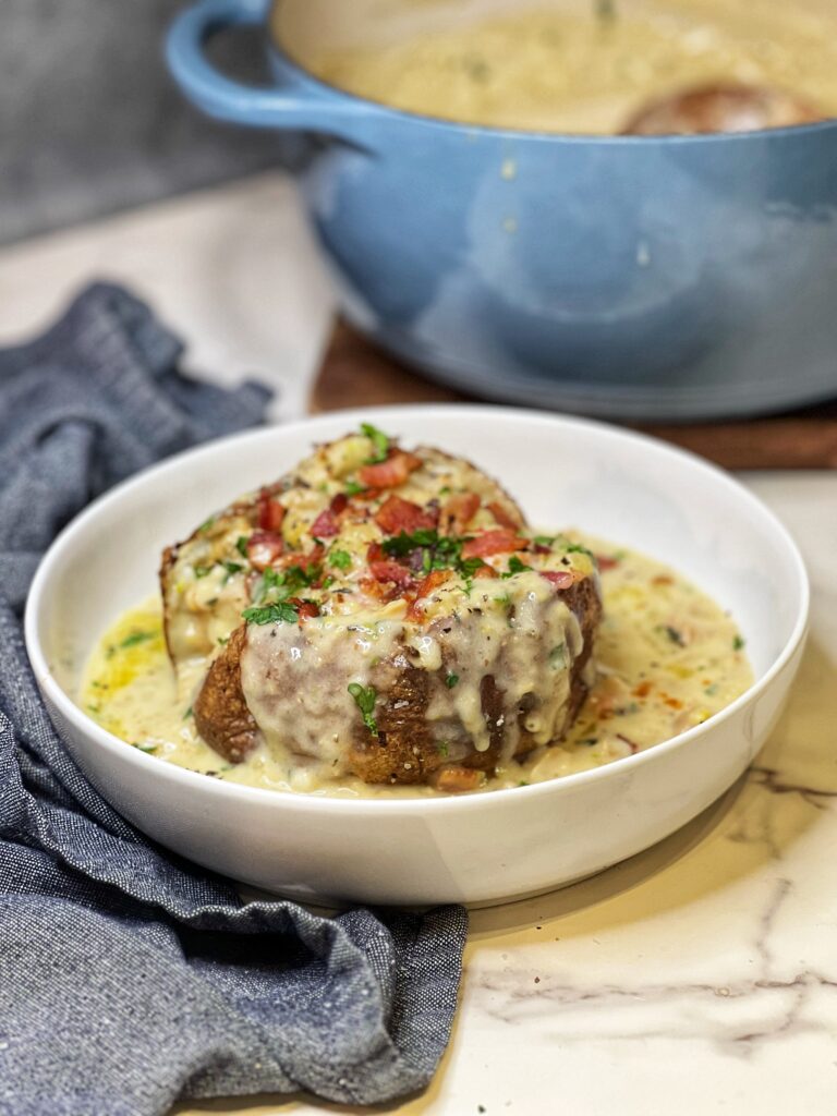 Creamy canned clam chowder poured on top of a baked potato that is sitting in a white bowl. The dish is garnished with crisp bacon and fresh parsley.