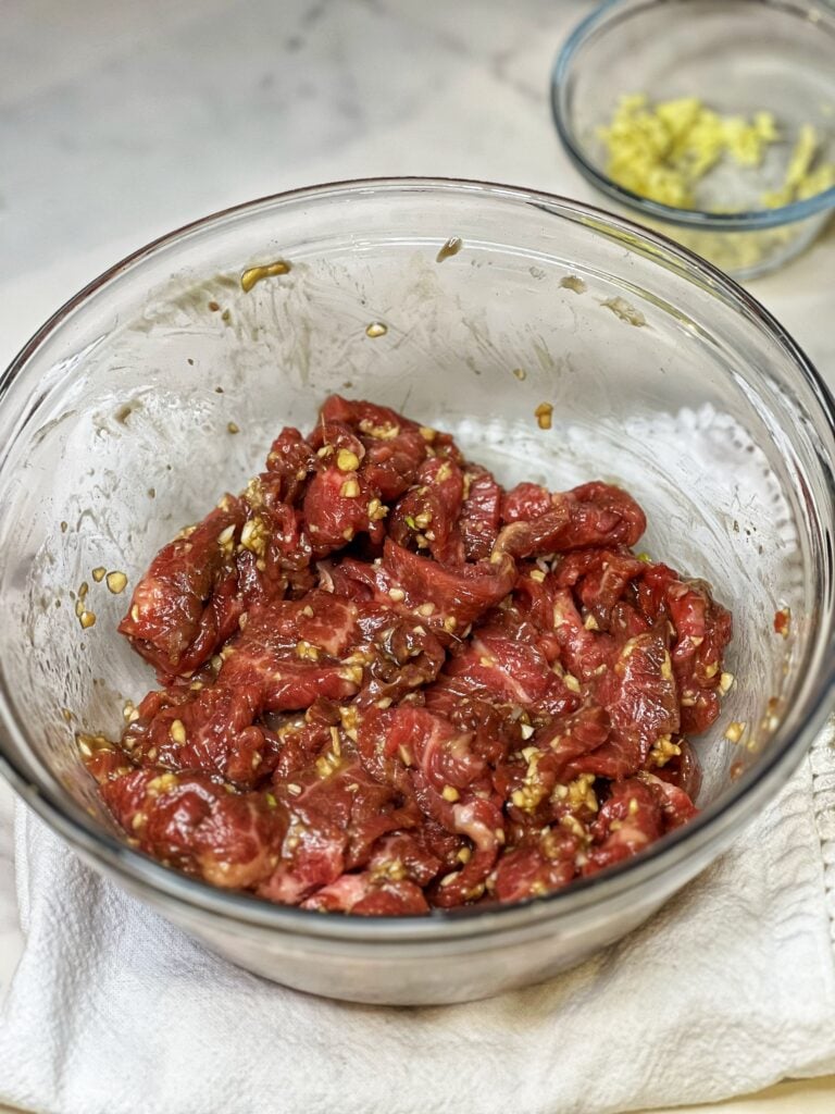 Thinly sliced flank steak in a glass bowl with a marinade of garlic, ginger and soy for Beef and Broccoli.