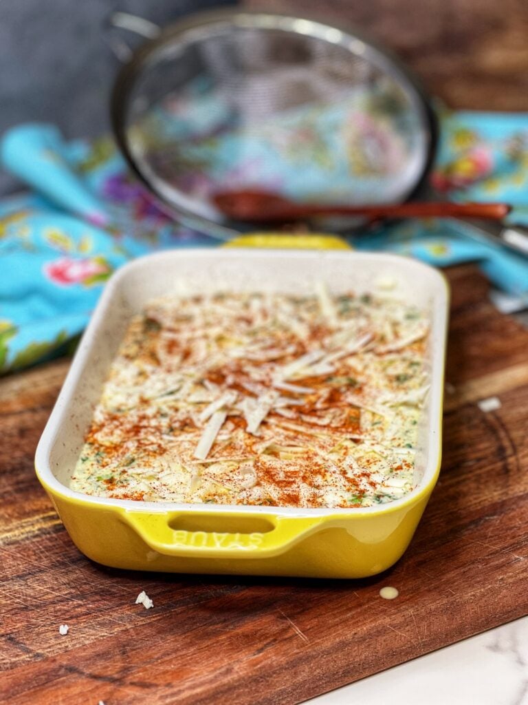 A small yellow, rectangular casserole dish filled with an egg and cottage cheese custard and topped with shredded white cheese and paprika. The dish is ready to be baked.