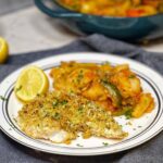 A white plate holding a filet of almond crusted baked wolffish with a side of stewed peppers, potatoes and tomatoes.