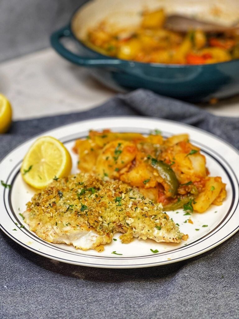 A white plate holding a filet of almond crusted baked wolffish with a side of stewed peppers, potatoes and tomatoes.