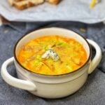 A white crock outlined in brown filled with Creamy Buffalo Chicken Soup. The soup is garnished with crispy tortilla strips, chopped celery leaves and blue cheese.