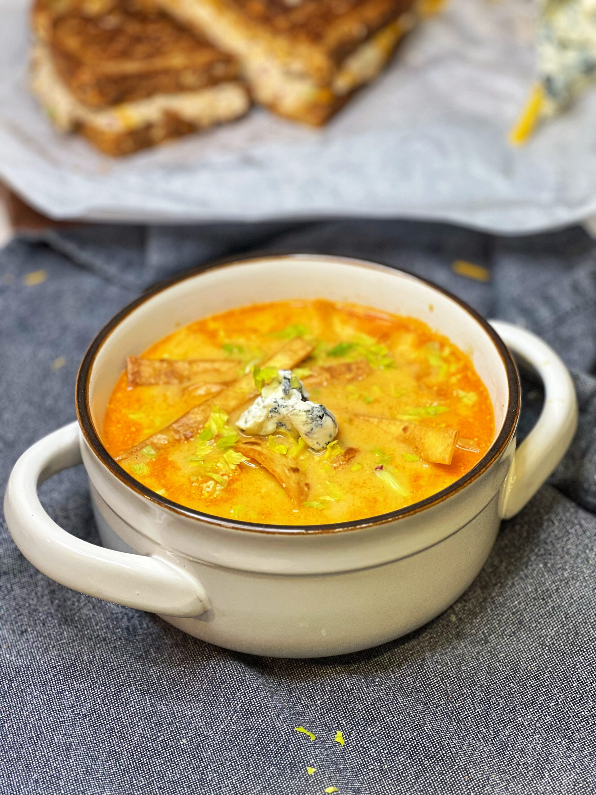 A white crock outlined in brown filled with Creamy Buffalo Chicken Soup. The soup is garnished with crispy tortilla strips, chopped celery leaves and blue cheese.