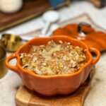 An orange, pumpkin-shaped baking dish filled with baked pumpkin bread pudding that is drizzled with a maple cream sauce.