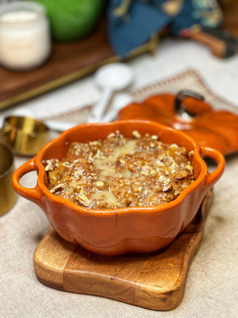 An orange, pumpkin-shaped baking dish filled with baked pumpkin bread pudding that is drizzled with a maple cream sauce.