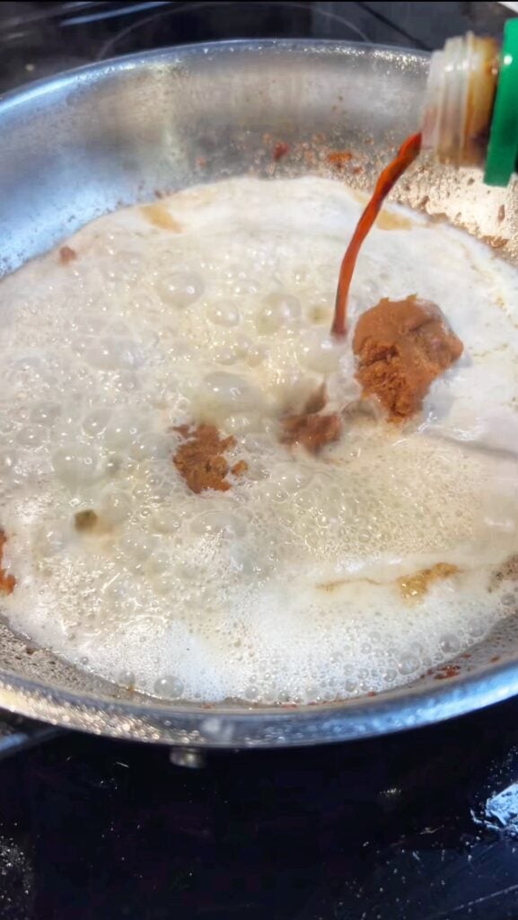 Pouring soy sauce into a pan with heated heavy cream and a spoonful of miso paste. 