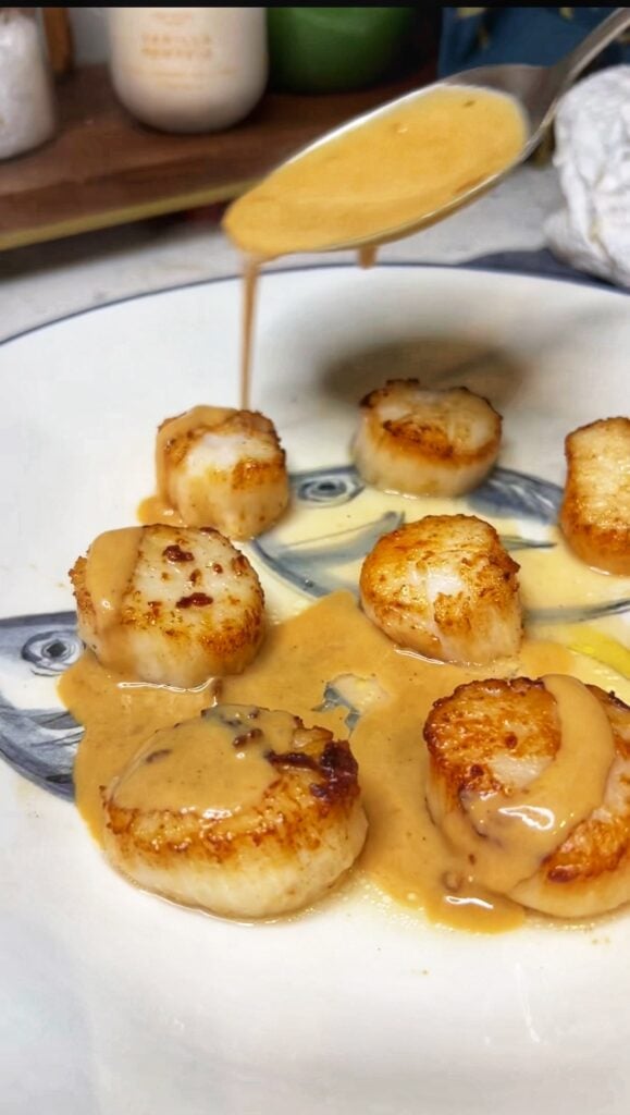 Spooning miso cream sauce over seared scallops. The sauce is brownish-orange in color.