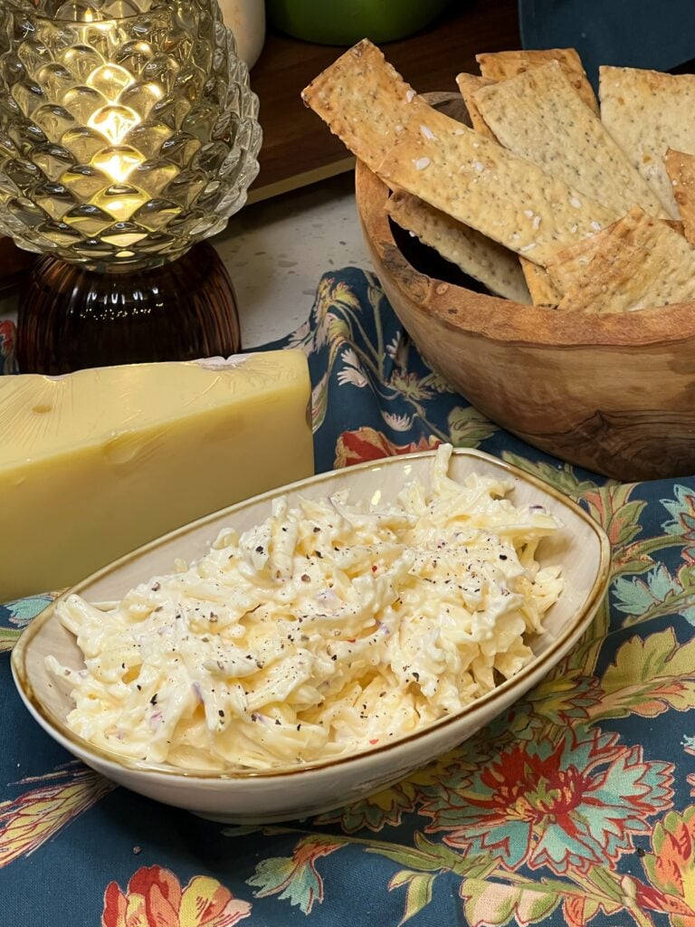 A tan, boat-shaped bowl filled with Jarlsberg Cheese Dip. The shredded cheese is mixed with a mayo-based sauce. The dip is garnished with cracked black pepper.